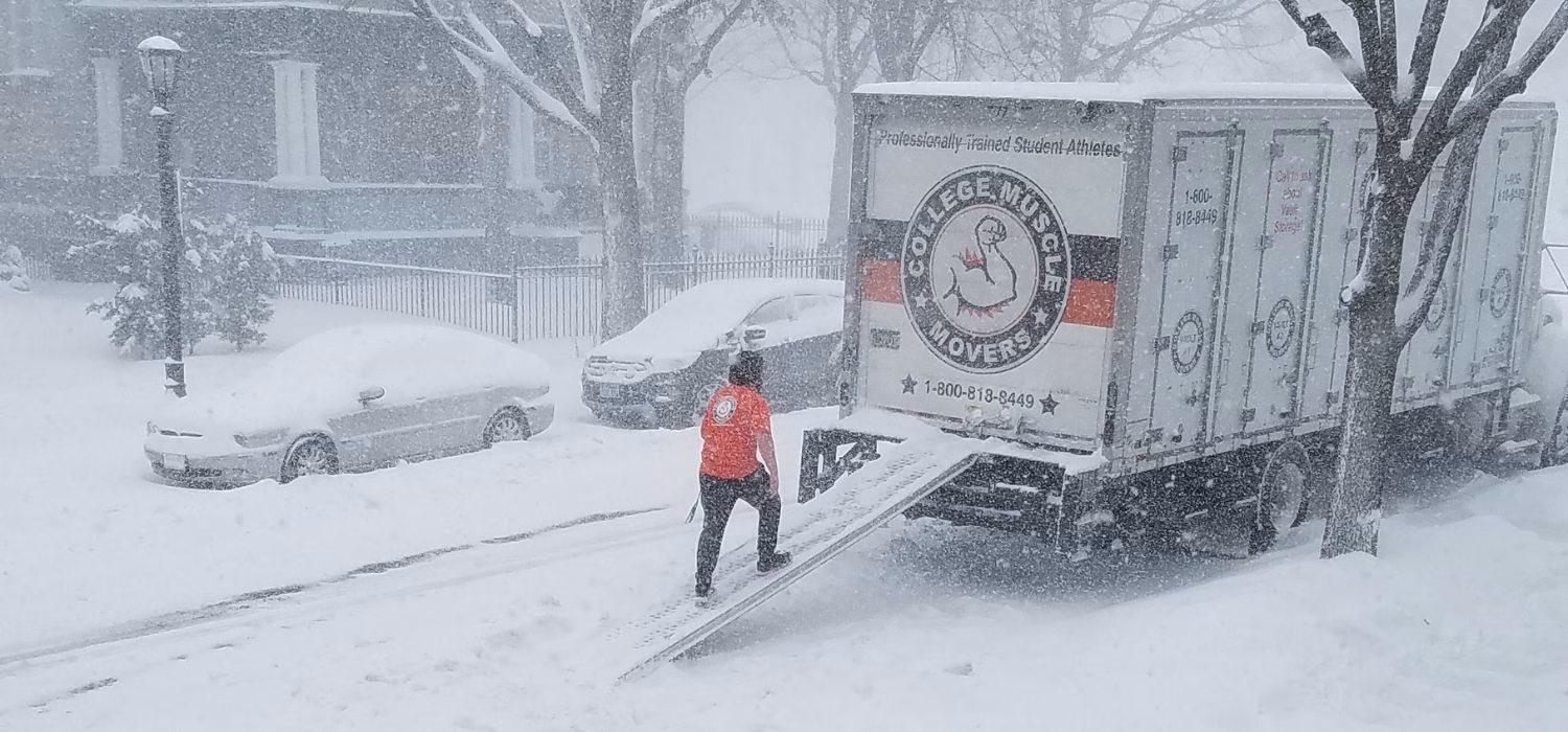 A mover walking up the ramp of a College Muscle Moves truck on a snowy residential street in Minneapolis, MN
