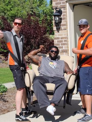 Two College Muscle Movers employees using shoulder straps to lift former Minnesota Vikings player Sharrif Floyd in an arm chair.